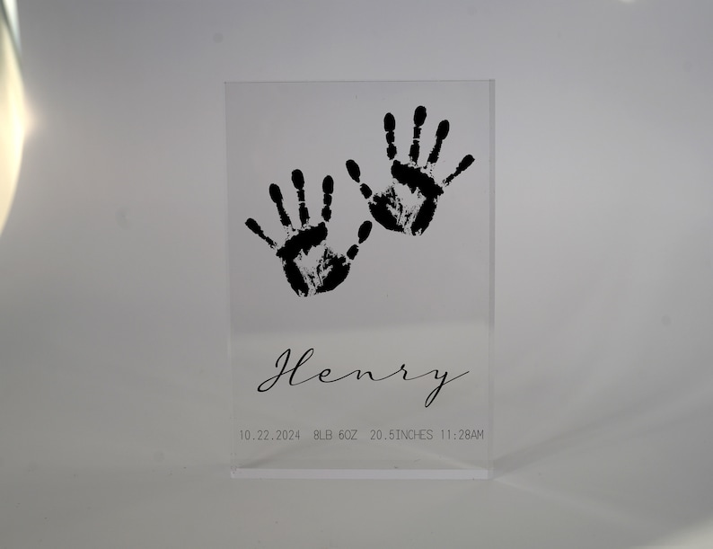 Op de afbeelding: Een heldere acrylblok met zwarte handafdrukken en de naam "Henry" gegraveerd. De datum "10.22.2024", het gewicht "8 LB 6OZ", de lengte "20.5 INCHES" en de tijd "11:28AM" zijn ook op de blok gedrukt.
