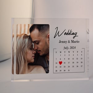 May include: A clear acrylic block with a photo of a couple and a calendar with the date July 15, 2024 highlighted. The text "Wedding" and "Jenny & Mario" are printed on the block.