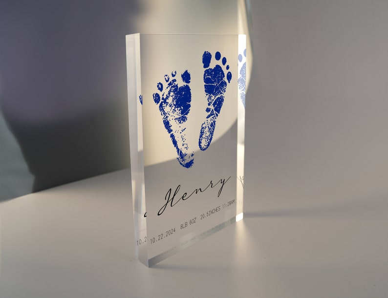 Op de afbeelding: Een transparante acrylblok met blauwe baby-voetafdrukken en de naam "Henry" gegraveerd. De datum "10.22.2024" en het gewicht en de lengte van de baby zijn ook opgenomen.