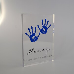 Op de afbeelding: Een transparant acrylblok met twee blauwe handafdrukken en de naam "Henry" in cursief. De datum "10.22.2024", het gewicht "8LB 6OZ", de lengte "20.5 INCHES" en de tijd "11:28AM" zijn ook op het blok gedrukt.