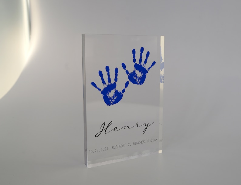Op de afbeelding: Een transparant acrylblok met twee blauwe handafdrukken en de naam "Henry" in cursief. De datum "10.22.2024", het gewicht "8LB 6OZ", de lengte "20.5 INCHES" en de tijd "11:28AM" zijn ook op het blok gedrukt.