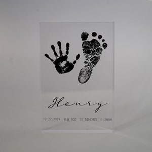 Op de afbeelding: Een heldere acrylblok met een zwarte handafdruk en voetafdruk, de naam "Henry", en de datum "10.22.2024", het gewicht "9lb 6oz", de lengte "20.5 inch", en de tijd "11:28AM".