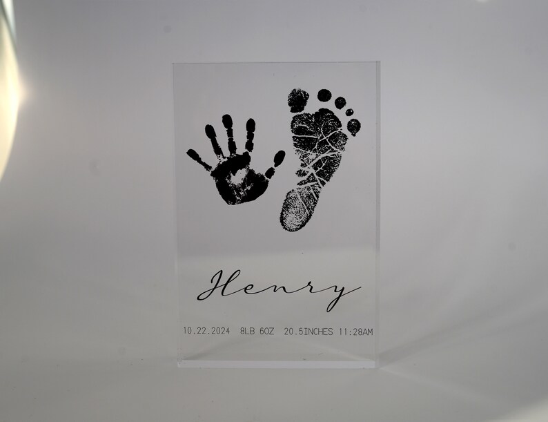 Op de afbeelding: Een heldere acrylblok met een zwarte handafdruk en voetafdruk, de naam "Henry", en de datum "10.22.2024", het gewicht "9lb 6oz", de lengte "20.5 inch", en de tijd "11:28AM".