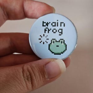Op de afbeelding: Een lichtblauwe button met de woorden "brain frog" in pixelachtige zwarte letters. Onder de tekst staat een groene pixelachtige kikker, met pixelachtige sterren links. De button is rond en wordt door een hand vastgehouden.