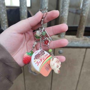 Può includere: Un portachiavi argentato con diversi ciondoli, tra cui una fragola rossa, una piccola statuina di gatto e un'etichetta a tema dessert con testo giapponese. L'etichetta presenta l'immagine di un dolce. Il portachiavi è tenuto in mano.
