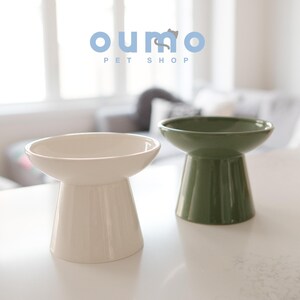 May include: Two elevated ceramic pet bowls, one white and one green. The bowls are on a white surface. The text "oumo PET SHOP" is visible in the background.