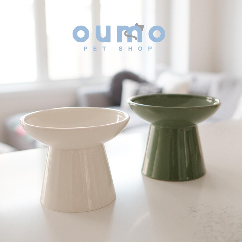 May include: Two elevated ceramic pet bowls, one white and one green. The bowls are on a white surface. The text "oumo PET SHOP" is visible in the background.