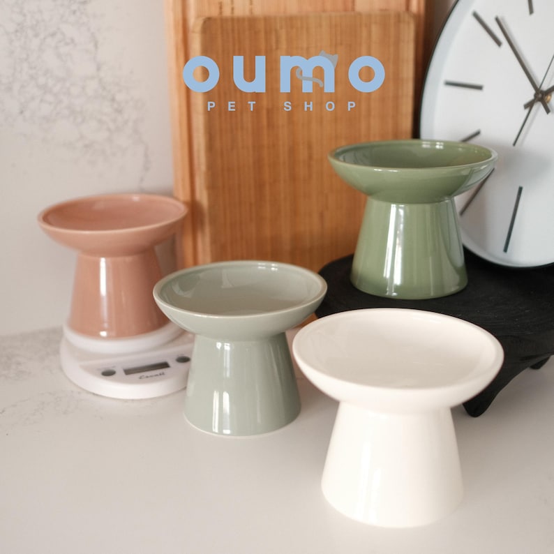 May include: Three elevated pet bowls in different colors: light brown, light green, and white. The bowls are elevated on a pedestal base. The text "oumo PET SHOP" is visible in the background.
