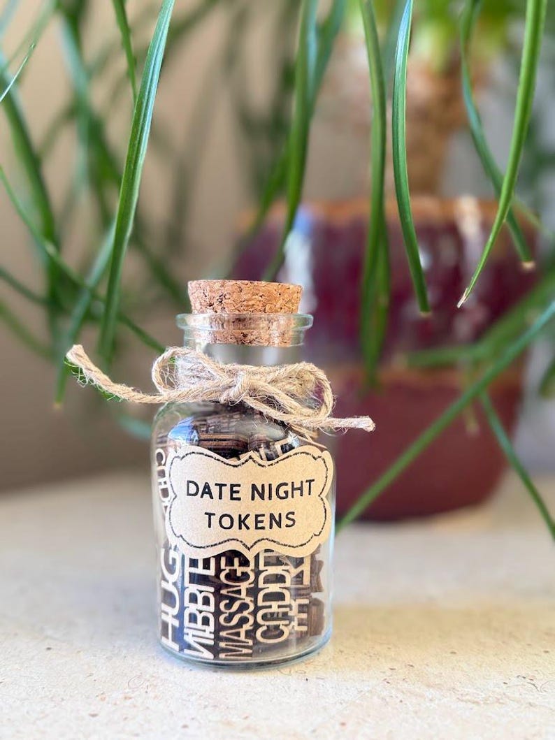 K&ouml;nnte beinhalten: Ein kleines Glas mit einem Korken und einer Schnur-Schleife, gef&uuml;llt mit Holzmarken. Auf dem Etikett steht "DATE NIGHT TOKENS". Die Marken tragen W&ouml;rter wie "HUG", "CUDDLE" und "MASSAGE".