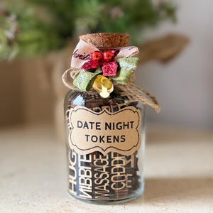 K&ouml;nnte beinhalten: Ein klares Glas mit Holzmarken, beschriftet mit "DATE NIGHT TOKENS". Das Glas ist mit einem Korken, einer dekorativen Schleife und Bindfaden verziert. Die Marken enthalten verschiedene romantische Aktivit&auml;tsvorschl&auml;ge.