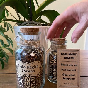 K&ouml;nnte beinhalten: Zwei Glasbeh&auml;lter mit Korken und Schnurakzenten, gef&uuml;llt mit Holzmarken. Ein Glas hat ein Etikett mit der Aufschrift "Date Night Tokens". Eine kleine Karte neben den Gl&auml;sern enth&auml;lt Anweisungen.