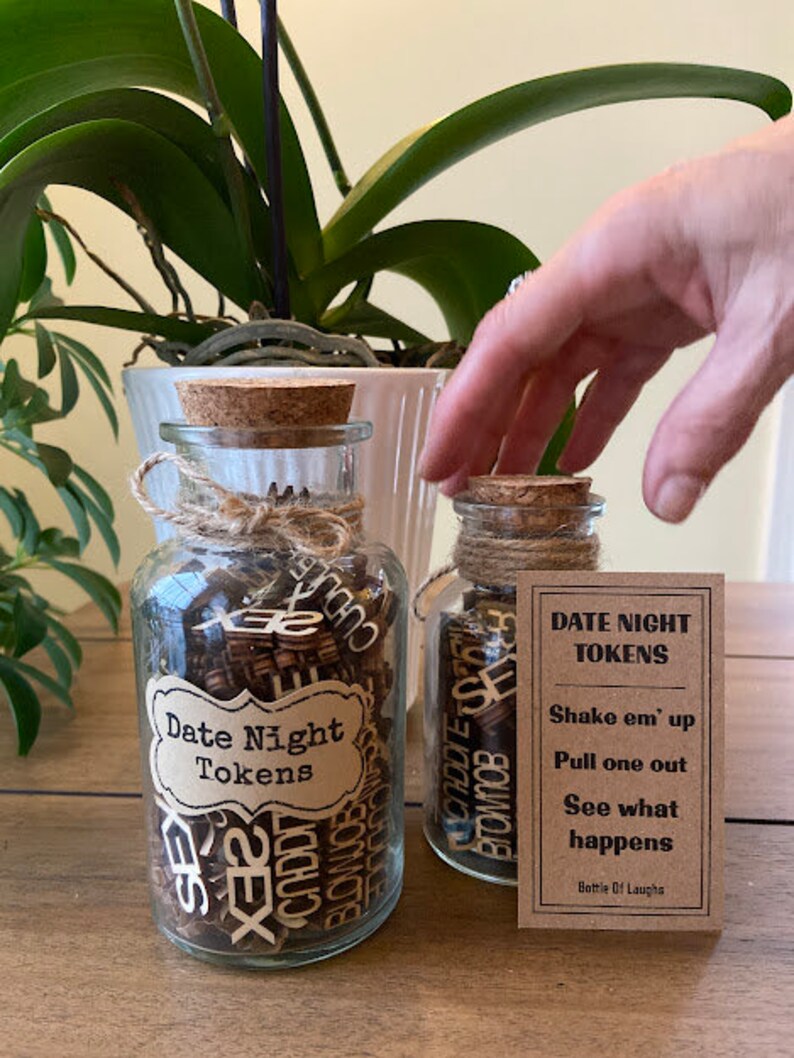 K&ouml;nnte beinhalten: Zwei Glasbeh&auml;lter mit Korken und Schnurakzenten, gef&uuml;llt mit Holzmarken. Ein Glas hat ein Etikett mit der Aufschrift "Date Night Tokens". Eine kleine Karte neben den Gl&auml;sern enth&auml;lt Anweisungen.
