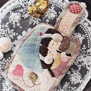 May include: A handmade quilted pouch with a handle, featuring an appliqué design of two figures embracing. The pouch has a pink heart and a red heart accent. The background is a white lace doily with a gold bird figurine.