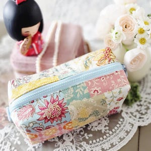May include: A rectangular pencil case with a patchwork design and a light blue zip. The case displays floral patterns in pink, yellow, and blue. A Japanese Kokeshi doll and a bouquet of white flowers are in the background.