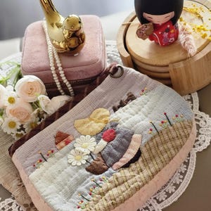 May include: An oval quilted pouch with a landscape design, featuring a yellow hat, daisies, and embroidery. The pouch has a pink border and sits on a lace doily. A gold bird ornament and a Japanese doll are also visible in the image.