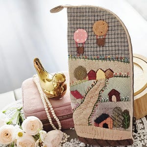 May include: A handcrafted, fabric pencil case with a whimsical landscape design. The case features a patchwork of various fabrics, depicting houses, trees, and hot air balloons. The case is approximately 8 inches tall and has a zipper closure.