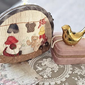 May include: A decorative cosmetic bag with a patchwork design featuring a clothesline scene and a teddy bear. A small, pink jewellery box with a gold bird ornament and a pearl necklace are also present. The items are on a white lace doily.