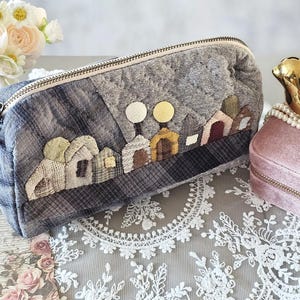 May include: A gray and blue quilted pencil case with a landscape design featuring small houses and two yellow circles. A gold bird figurine sits atop a pink velvet jewelry box, with a pearl necklace draped around it.