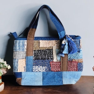 May include: A patchwork tote bag with a variety of blue, brown, and beige fabric squares. The bag has two handles and a small blue tassel with a fish charm. The bag is sitting on a wooden surface.