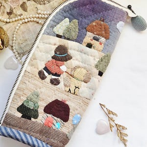 Może przedstawiać: Pikowane piórnik z fantazyjnym krajobrazem. Etui przedstawia scenę z osobą spacerującą z psem, drzewami i domem. Etui posiada zapięcie na zamek błyskawiczny i materiałowy frędzel. Paleta kolorów obejmuje odcienie niebieskiego, brązowego i zielonego.