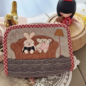 Puede incluir: Un estuche acolchado con una escena caprichosa de un conejito y un gato en un sofá marrón, con una lámpara. El estuche tiene un borde a cuadros rojo y blanco y una base estampada gris. Una figura dorada y una muñeca japonesa están en el fondo.