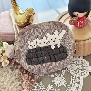 May include: A round, brown fabric pouch with a cat applique. The pouch features three white cats peeking over a dark brown, textured border. The background includes a gold bird figurine, a pink jewelry box, and a Japanese doll.