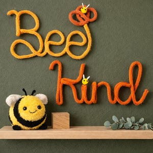 May include: Wall decor featuring the words Bee Kind spelled out in orange and yellow knitted yarn script. Two small yellow and white bee accents are attached to the letters. A plush yellow and black bee toy sits on a wooden shelf below.