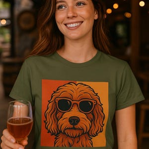 May include: Olive green t-shirt with a square graphic of a cartoon dog wearing sunglasses. The dog is brown with black sunglasses, set against an orange and yellow background. The person is holding a glass of amber liquid.