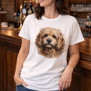 May include: White t-shirt with a watercolour illustration of a Cavapoo dog. The dog has brown and tan fur and is centered on the shirt. The person is wearing blue jeans and is leaning against a wooden bar.