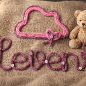 May include: A knitted purple yarn sign spelling the word Leven in cursive script, decorated with small white flowers and a heart shape. Above the text is a pink and purple knitted cloud shape with a heart, accompanied by a tan plush teddy bear.
