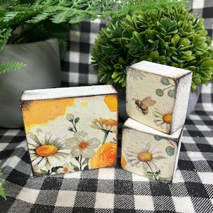 May include: Three decorative wooden blocks with floral and bee designs. The blocks feature daisies, roses, and a bee, with a distressed white border. The background includes greenery and a black and white checkered pattern.