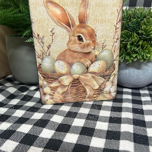 May include: A rectangular wooden sign featuring a brown bunny in a basket with speckled eggs. The bunny has long ears and is surrounded by sprigs and a bow. The background is a beige color with faded text.