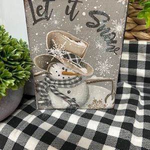 May include: A wooden block with a snowman illustration wearing a cowboy hat and a black and white checkered scarf. The block has the words "Let it Snow" in black script on a gray background with snowflakes.