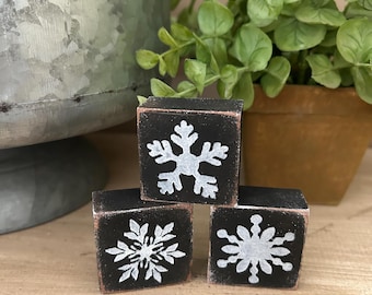 May include: Three small, black wooden blocks with white snowflake designs. The blocks are arranged on a wooden surface, with a blurred background featuring greenery and a metal container. The snowflake designs vary slightly.