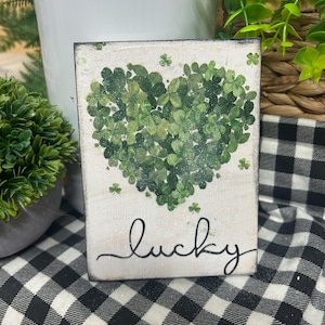 May include: A small, rectangular wooden sign with a heart-shaped design made of green shamrocks. The word "lucky" is written in cursive below the heart. The background is white with a distressed finish, and the sign is placed on a black and white checkered cloth.