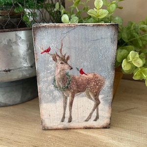 May include: A rustic wooden block features a winter scene with a deer wearing a wreath and two red cardinals. The deer has brown fur with white spots. The background is a snowy, light blue. The block has a distressed, aged appearance.