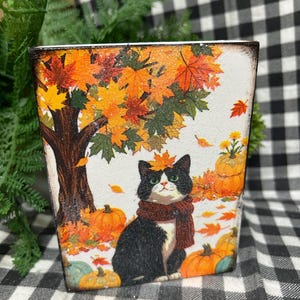 May include: A rectangular wooden block featuring a printed autumn scene with a black and white cat wearing a brown scarf, sitting under a tree with orange and yellow maple leaves, surrounded by orange pumpkins on a white background with glitter.