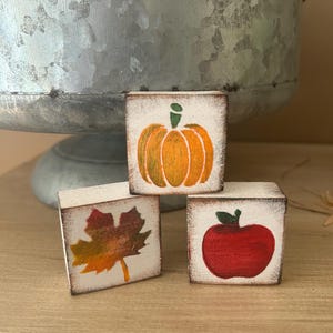 May include: Three small, square wooden blocks with fall-themed designs. One block features an orange pumpkin, another a red apple, and the third a colorful autumn leaf. The blocks have a distressed, rustic look.
