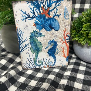 May include: A rectangular decorative block featuring a marine life design. The artwork includes blue seahorses, coral, and a seashell against a neutral background. The block is accented with blue, orange, and green elements, evoking a coastal theme.