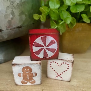 May include: Three small, hand-painted wooden blocks. One is red with a white peppermint design, another is white with a gingerbread man, and the last is white with a candy cane heart. The blocks are rustic and decorative.