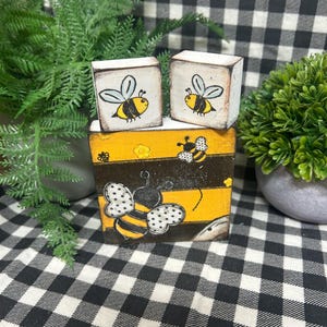 May include: Hand-painted wooden blocks featuring bee designs. The set includes two small blocks with bee illustrations and a larger block with a bee and honeycomb pattern. The color scheme includes yellow, black, and white.