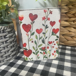 May include: A small, rectangular container with a white background, decorated with red heart-shaped flowers and green leaves. The container has a distressed, vintage look, and is placed on a black and white checkered surface.