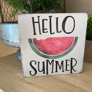 May include: A square, white wooden sign with the words "HELLO SUMMER" in black lettering. A red watermelon slice with a bite taken out of it is in the center. The sign has a distressed, rustic appearance.