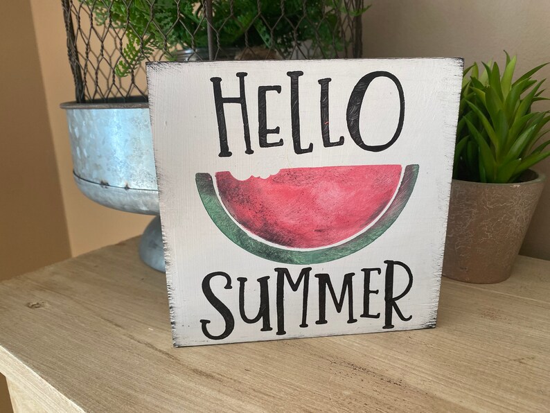 May include: A square, white wooden sign with the words "HELLO SUMMER" in black lettering. A red watermelon slice with a bite taken out of it is in the center. The sign has a distressed, rustic appearance.