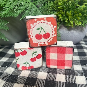 May include: Three small, square wooden blocks with cherry designs. One block features a red and white gingham pattern, another has a white background with red cherries, and the third has a red background with a white cherry design.