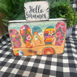 May include: A wooden block sign with the words "Hello Summer" on top of a rectangular box. The box features a beach scene with colorful flip-flops, a beach ball, sunglasses, and a watermelon float.