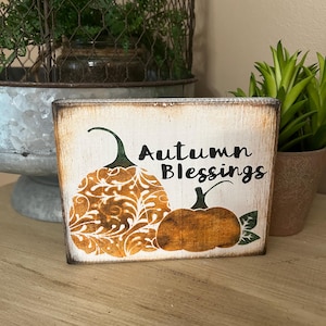 May include: A rustic wooden sign with the words "Autumn Blessings" in black script. The sign features two orange pumpkins with green stems and leaves. One pumpkin has a decorative pattern. The background is a distressed white.