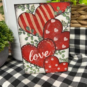May include: A rectangular decorative piece featuring red hearts with various patterns, including stripes and polka dots, and the word "love". Green leafy accents surround the hearts. The background is white with a distressed look.