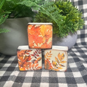 May include: Three small square wooden blocks featuring a decorative autumn leaf pattern. The blocks are arranged in a pyramid stack, with one block on top of two others. Each block displays orange, yellow, and brown foliage illustrations.