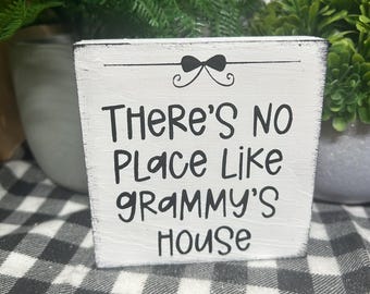 May include: A square white wooden sign with distressed black edges featuring a black bow graphic at the top and the black text "There's NO PLacE LiKe gRaMMY's HouSe" printed in a mix of uppercase and lowercase letters on the front surface.